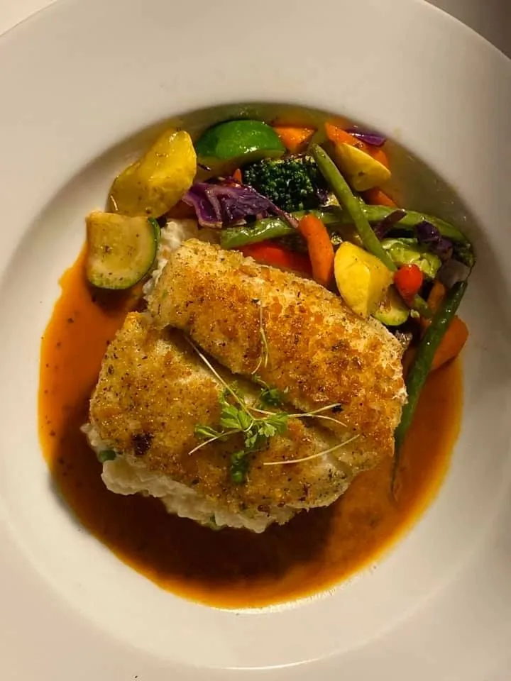 A crusted halibut fillet over a golden sauce, topped with roasted vegetables and microgreens